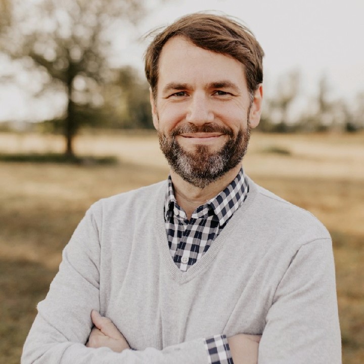 Josh Fleig a man in a grey sweater and arms crossed smiles. He is outside. 