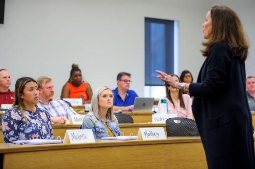 Woman in black dress teaches in front of classroom of onlooking students. 